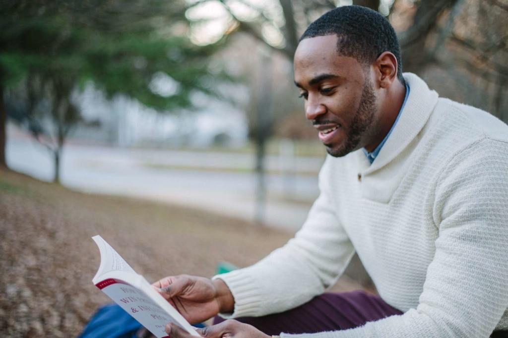 A man sitting near the river, holding a book and reading attentively, symbolizing the act of engaging in leisurely reading for knowledge or enjoyment